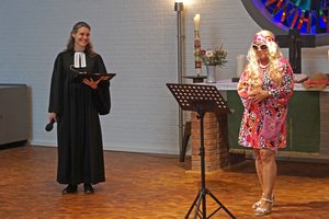 Eine Frau in geistlicher Kleidung mit einer aufgeschlagenen Mappe in der Hand, eine weitere bunt gekleidete Frau steht vor einem Notenständer
